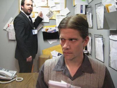 In the foreground, a nervous man holds some papers while sitting at his desk in a cluttered office. Behind him, a bearded man in a suit with a badge stands, speaking.