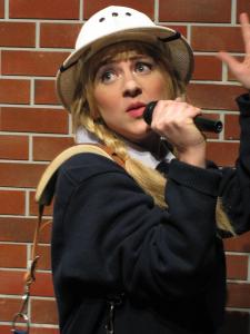 A postal worker with blond braids sings into a microphone in front of a brick wall.