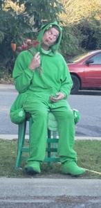 A person in a bright green grasshopper costume is sitting on a green chair, chewing on a some fake flowers. Behind is the street with a red car and some green foliage.