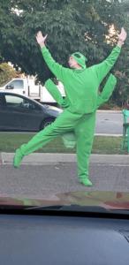 A picture from inside of a car. At the bottom of the picture is the dashboard. In the parking lot, a person in a bright green grasshopper costume is dancing, all four arms up and one leg kicked to the side. In the distance is the street with cars and trees.