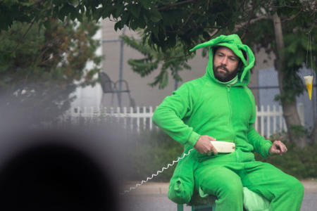 A bearded man in a bright green grasshopper costume sits, looking serious. He is holding a phone receiver to his belly. In the distance are trees, a white picket fence, and patio furniture. In the foreground, something out-of-focus obscures the bottom left corner.