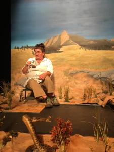 A woman in a white shirt sits on a very short stool looking into her coffee cup as though it has something gross in it. She is surrounded by an unfinished prairie floor, including an angry taxidermied rattlesnake in the foreground. Behind is a painted backdrop with a distant mountain.
