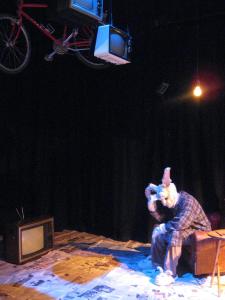 A large rabbit wearing a ratty robe and bunny slippers slumps in a lazy boy recliner. There is a television on the floor infornt of him and two more hanging above him. The floor is covered with newspaper.