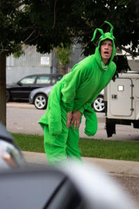 A picture from inside of a car. In the parking lot, a person in a bright green grasshopper costume is dancing, bent over with their hands on their knees. In the distance is the street with cars, buildings, and trees. In the foreground, something out-of-focus obscures the bottom left corner.