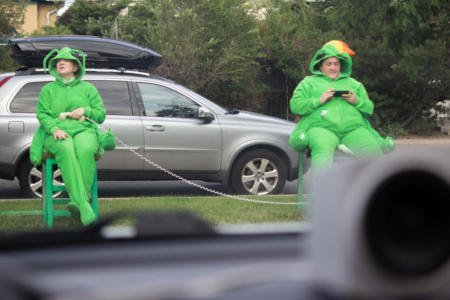 A picture from inside of a car. At the bottom is the dashboard with an out-of-focus speaker sitting on it. In the distance, two people in bright green grasshopper costumes are sitting on green chairs. One has a phone receiver held to their belly. The other is looking at a cell phone. Behind them is a sliver car parked on the street.