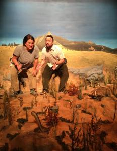 Two people are standing, knees bent in action. The woman is angrily talking, the man looks confused and surprised. They are standing a dry prairie scene, where a taxidermied badger and rattlesnake are facing off. The backdrop is a painted scene with distant mountains.