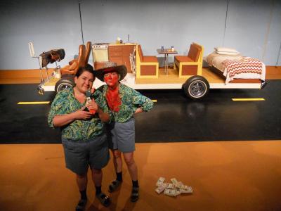 Two people in matching outfits stand next to a pile of money. One holds a small wooden duck, the other has bright red powder covering her face and shirt. In the background, on a painted road, is the insides of an RV sitting on a platform with wheels.