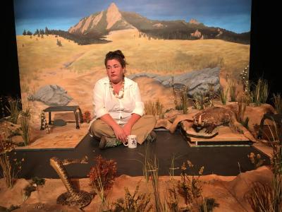 A woman in a white shirt sits cross-legged on the floor, looking despondent. She has a coffee cup in front of her. She is surrounded by an unfinished prairie scene, including a taxidermied badger facing off with a taxidermied rattlesnake. Behind all of them is a painted backdrop with a distant mountain.