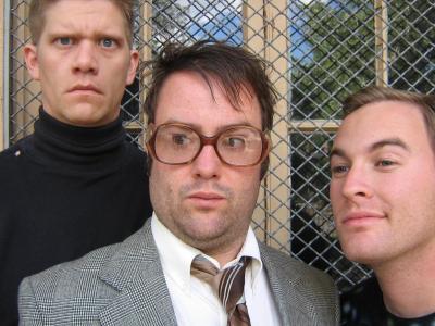 Close-up of three awkward men in front of a window grate. On the left is an intense man with a buzz cut and black turtleneck. In the center, a man in a suit and large glasses purses his lips. On the right is a smiling man with a space t-shirt on