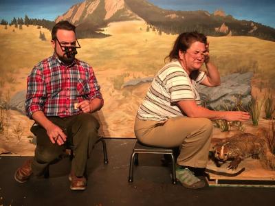 A bearded man in a plaid shirt is sitting on short stool. He is wearing magnifying glasses. Next to him is a woman, also sitting on a short stool. She is wearing glasses and is rubbing her eyes, annoyed. She if facing sideways. Under her outstretched hand is an angry taxidermied badger. Behind all of them is a painted backdrop with a distant mountain.