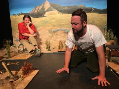 A bearded man is on all fours. He looks concerned and seems to be facing off with a taxidermied rattlesnake. They are surrounded by a partially finished prairie floor. In the background a woman in a bright orange shirt sits on a small stool looking on, angry or bored or both. Behind all of them is a painted backdrop with a distant mountain.
