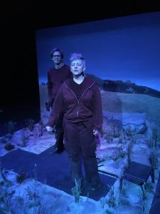 Two people wearing black stand in a partially finished scene of a prairie with distant mountains. Some of the flooring is bare. One person talks with their eyes closed as the other stands behind, looking at them. Everything is bathed in blue light.
