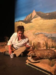 A woman in a white top sits cross-legged on the floor with a coffee cup. She is talking to a taxidermied angry badger. Behind them is a painted prairie scene with a distant mountain.