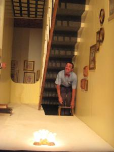 A man stands on top of a ladder in an upside down stairwell. There are pictures on the wall that are upside down and there is a ceiling light fixture on the floor. 