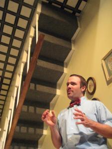 A man is standing and talking. There is a upside-down staircase above him. 