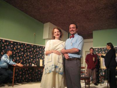 In the foreground, a couple holds each other while smiling in a strained way. She is pregnant and he wears a bow tie. In the background, three other people argue. Strangely, they are all standing in a room that is upside down. Pink carpet covers the ceiling.