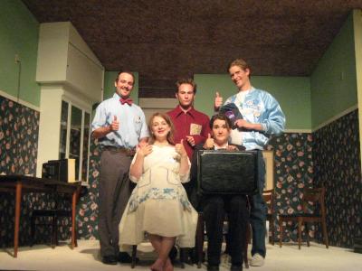 Four smiling people give the camera a thumbs up while the man in the middle looks annoyed. They are grouped together in the middle of an upside down room.