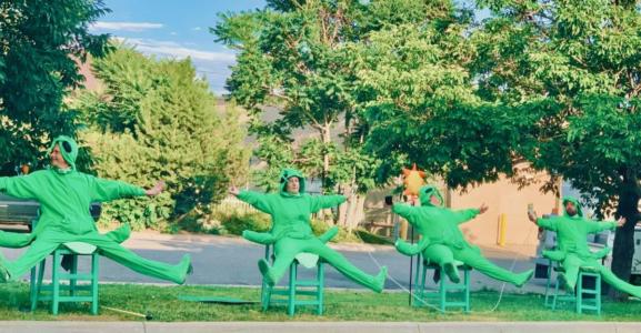 Four people in bright green grasshopper costumes sit outside in green chairs. They all have their arms and legs stretched out. In the distance is the street with cars, buildings, and trees.