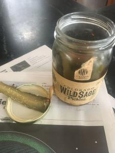 On a table sits an open jar of pickles on top of a newpaper. to the side of the jar is the lid, with a single pickle spear on it. 