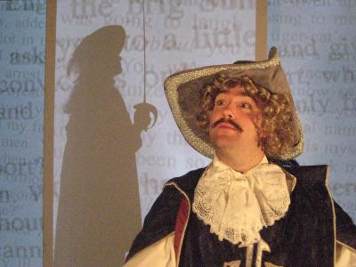 A man with a mustache and curly blond hair stands regally in a musketeer outfit. In the background there is a silhouette of another musketeer holding a sword, surrounded by projected text.