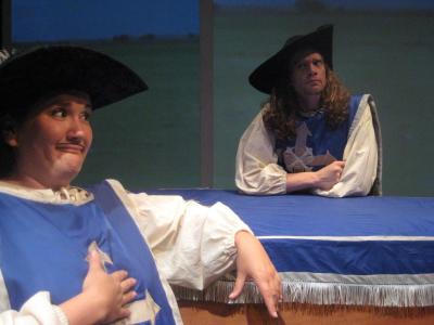 Two people dressed as musketeers lean on a coffin draped in a blue and white cloth. The musketeer in the foreground has a mustache drawn on her face.