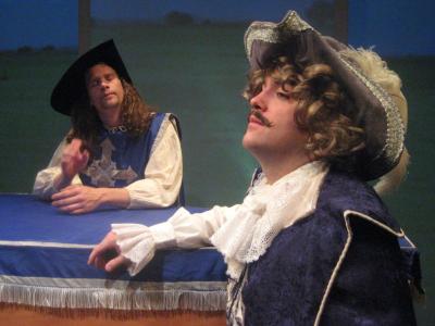 Two people dressed as musketeers lean on a coffin draped in a blue and white cloth. 