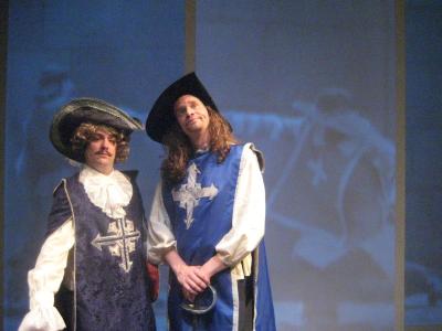 Two men dressed as musketeers pose in front of a giant screen that has video of musketeers fighting projected onto it.