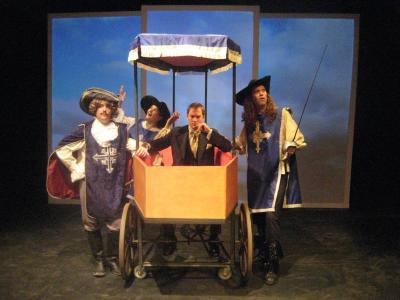 A man sits in a carriage that is shaped like a coffin. Surrounding him are three people dressed as musketeers. Behind them, an image of the sky is projected on a giant screen.