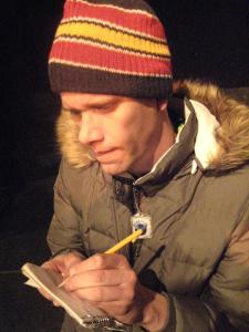 A man in a parka and knit cap bites his lip in concentration as he writes in a small notebook. 