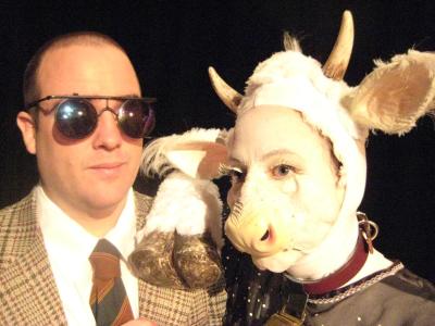 Close-up of a man in dark glasses and a woman dressed as a white cow. She has her hoof resting on his shoulder.