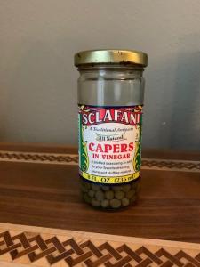A half full jar of capers on a wooden surface with interwoven inlay. 