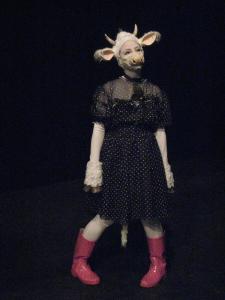 A woman dressed as a white cow in a polka dot dress and pink rain boots stands, surrounded by darkness.