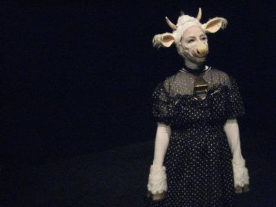 A woman dressed as a white cow in a polka dot dress stands, surrounded by darkness.