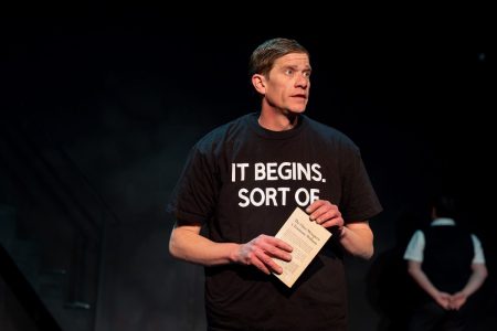 Close-up of a man speaking. He holds a copy of The Glass Menagerie and looks like he is wearing too many t-shirts. The top one says It begins. Sort of. In the distance is the back of another person facing upstage.