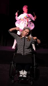 A woman in a wheelchair is making a funny face, with pursed lips. She is wearing black and white and is holding her pointer fingers up towards each other. Behind her, on a platform is a person in a white body suit with an inflatable rainbow unicorn pool float around their body. They have a child's xylophone around their next and are playing it with little wooden mallets.