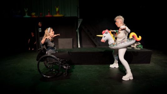 Two people face off, their hands up to the sides and they are both talking in a comically threatening way. One of them is in a wheelchair and wears black and white. The other wears a white body suit with an inflatable rainbow unicorn pool float around their body. In the background there are two people not paying attention to the center stage ruckus.