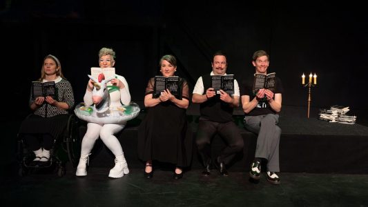Five people sit, looking at scripts of The Glass Menagerie. On the left is a woman in a wheelchair, wearing black and white. Next to her is a person in a white bodysuit with an inflatable rainbow unicorn pool float around their body. Then is a dark-haired woman in a balcony dress, followed by a mustached man in a white shirt and black sweater vest. Finally there is a man in grey pants and a t-shirt with something unintelligible printed on it. To the side is a lit candelabra on a stand and a pile of more scripts. There is blackness all around them.
