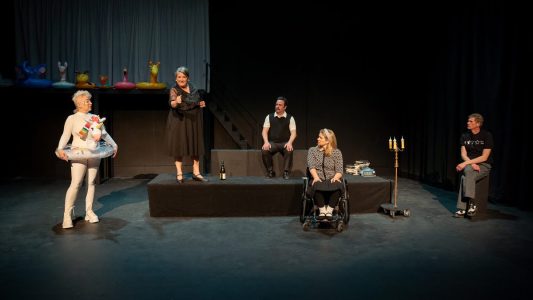 There are five performers on a black stage, with a long black platform in the middle of it. To the left is a person dressed in a white body suit with an inflatable rainbow unicorn pool float around their body. On the platform is a dark-haired woman in a black dress. She is talking and smiling. Sitting in the middle of the platform is a mustached man in a white shirt and black sweater vest. In front of the platform is a woman in a wheelchair, turned to listen to the speaker. She wears black and white clothing. To the side is a man seated on a box, wearing grey pants and a black t-shirt with white stars on it. In the deep background there are some colorful animal  pool floaties.