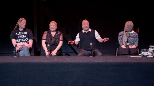 Four people sit behind a platform. They all have pantyhose on their heads. The first wears a t-shirt that says something about a lesson, but it is not all in view. The second is in a black dress. The third is talking with his hands out and wears a white shirt and a black sweater vest. The fourth wears black and white and is in a wheelchair. They all have microphones in front of them. There is a pile of books on the edge of the platform.