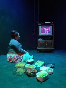 A person sits on the ground watching television. Next to her are many casseroles.
