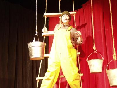 A sailor wearing bright yellow overalls and a pipe stands on a rope ladder surrounded by metal buckets hanging from rope. In the background is a red curtain.