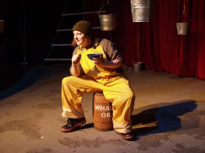 A sailor wearing bright yellow overalls and boots sits on a barrel marked “whale oil” while holding a spoon and a metal bowl. In the background is a red curtain, a rope ladder, and several hanging buckets.