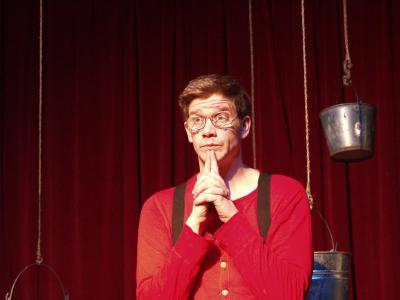 A man in a red top, black suspenders, and glasses looks out inquisitively. Behind him are several hanging buckets in front of a red curtain.