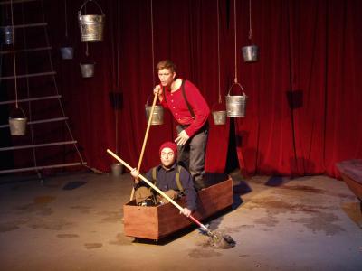 Two men, one seated and one standing, are “rowing” in a wooden coffin, using mops as oars. In the background is a red curtain, a rope ladder, and several hanging buckets.