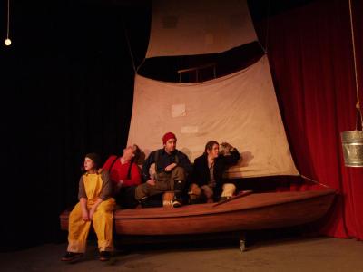 Three sailors sit in a small sailboat. A fourth sits on the edge of the boat, wearing bright yellow waders