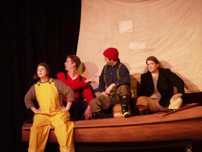Three sailors sit in a small sailboat. A fourth sits on the edge of the boat, wearing bright yellow waders