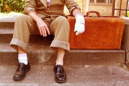 Shoulders down image of a man in a brown suit, he has one sock on his foot and one sock on his hand. He is resting on a leather suitcase. 