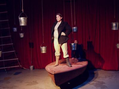 A woman stands on the front of a boat. she is wearing a peacoat, one boot and one sock that looks like a wooden leg. in the background there is a red curtain and several metal buckets hang from above. A man hold the back of the boat.