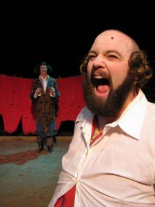 In the foreground, a bald man with a big beard and a beauty mark on his forehead is yelling. In the background, a man in a patterned jacket stands in a shallow mud pit, smiling. A clothing line of red long underwear hangs behind.
