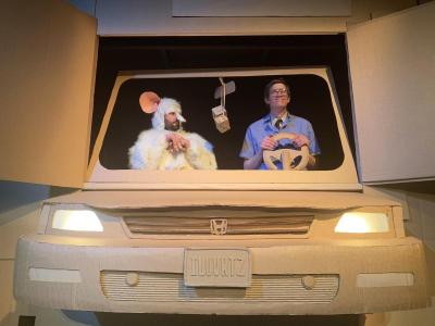 A lifesize car made out of cardboard with a “I love Rats” license plate is facing the camera.  In the drivers seat is dorky looking man wearing a blue jumper with a patch of a rat on his pocket. Next to him sits a giant white rat who is looking at the man a bit concerned.  The man is smiling and there are cardboard dice hanging from the cardboard rearview mirror.